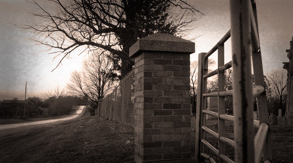 A fenced, brick-walled entrance gate stands beside Bloods Point Road, a rural Illinois road lined with trees in a sepia-toned image.