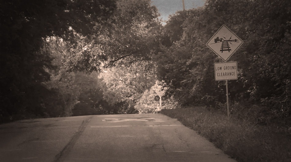 A rural road with a "Low Ground Clearance" sign, indicating a railroad crossing ahead on Haunted Munge Road. Dense trees and foliage surround the area, adding an eerie atmosphere to the journey.