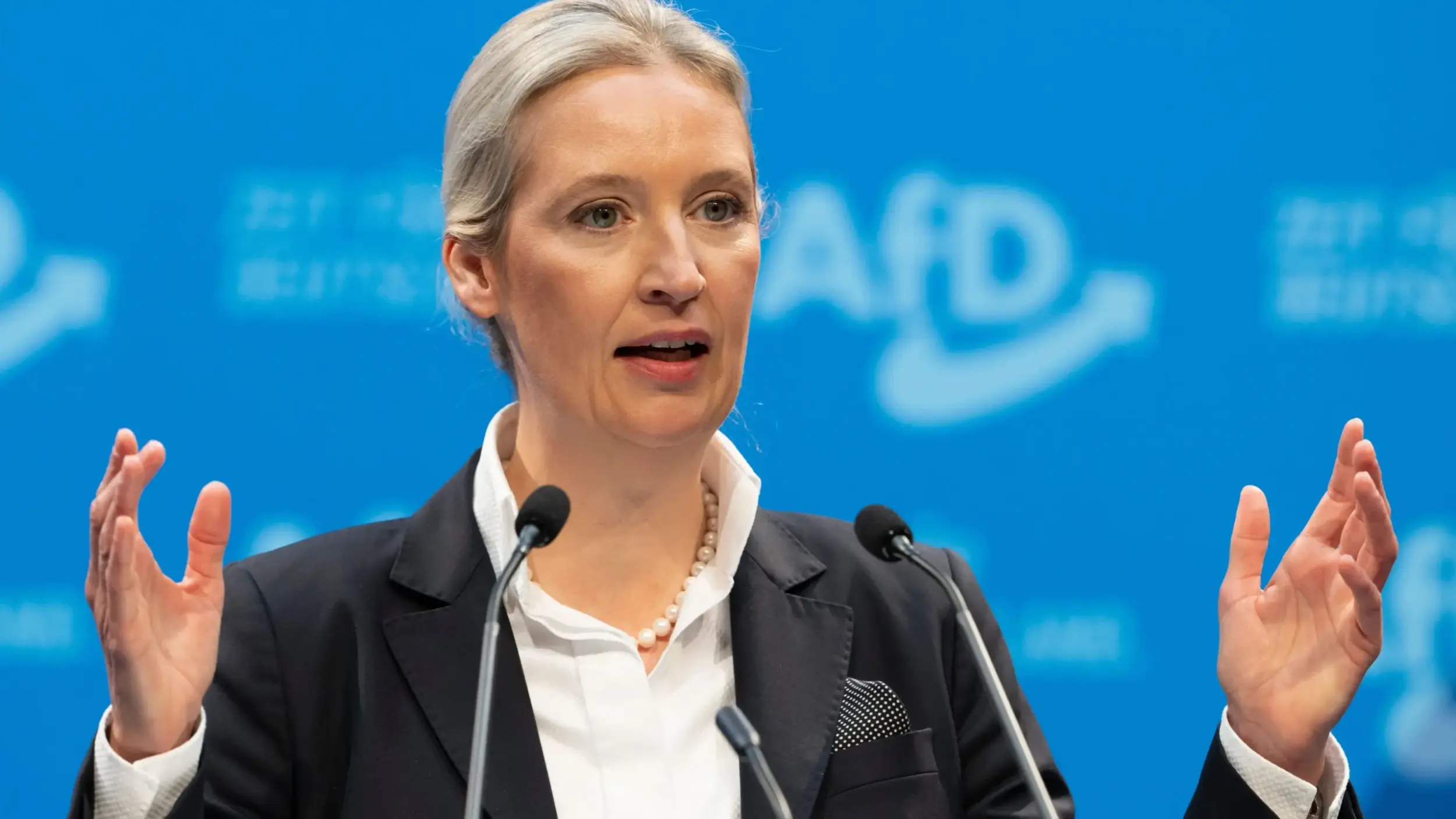 Politician speaking at a conference with microphones and a blue background, emphasizing key points during her address.