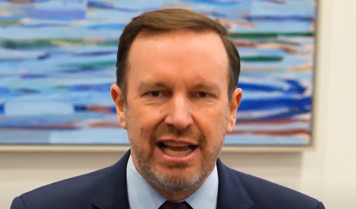 Man in a suit speaking passionately in front of an abstract blue painting, conveying a message with a focused expression.