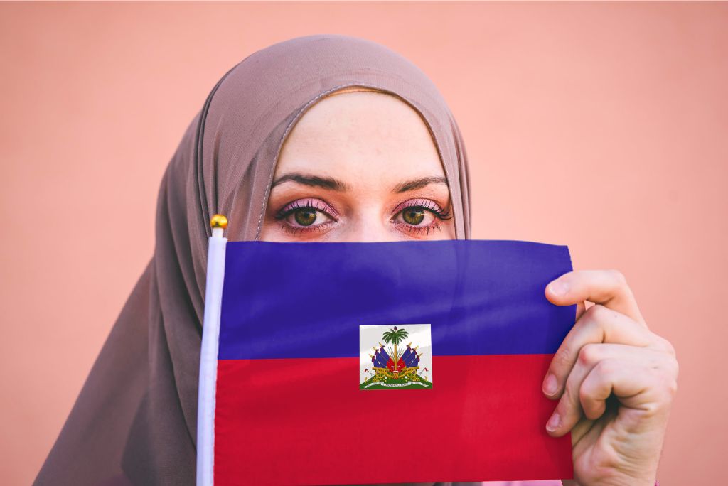 Woman in a hijab holding the Haitian flag close to her face, symbolizing cultural identity and pride against a soft pink background.