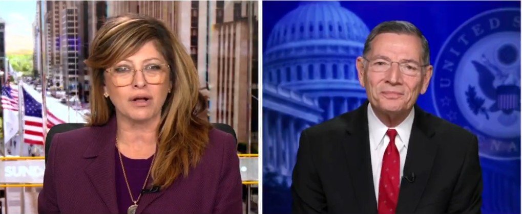 News interview featuring a female host and male guest discussing current political issues, with a backdrop of the U.S. Capitol and American flags.