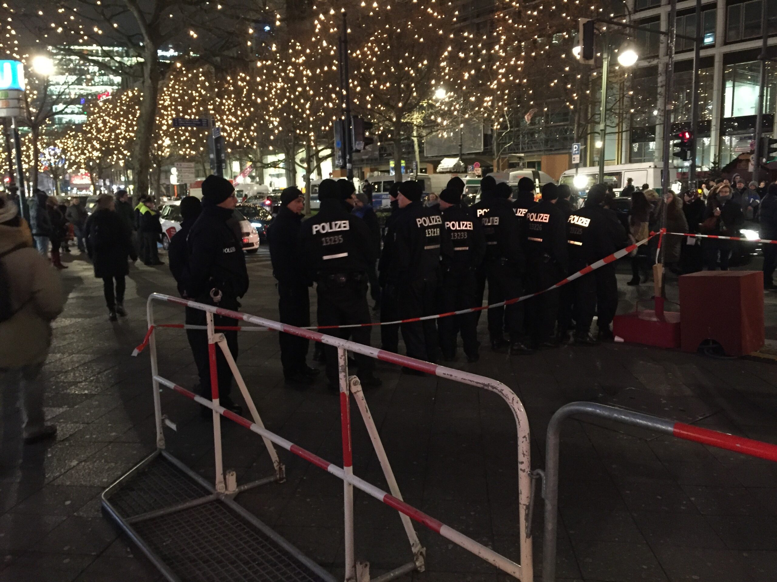 Police officers in uniform stand behind barriers at a crowded urban event, with festive lights illuminating the scene.