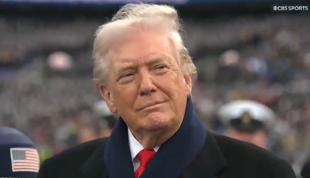 Former President Donald Trump smiles while attending a public event, dressed in a black coat and blue scarf against a blurred crowd background.