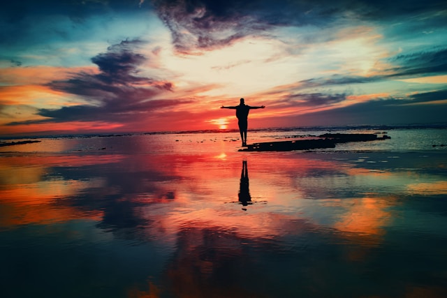 A man standing in shallow water, looking at the sun during dusk with open arms.