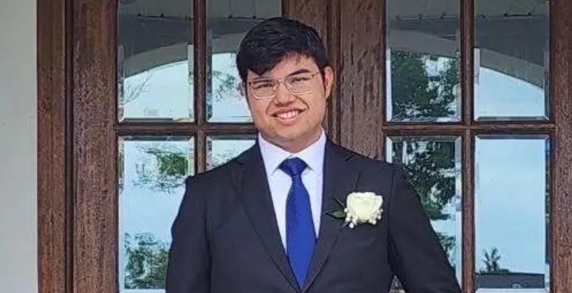 Young man in a formal suit with a blue tie and white boutonniere, smiling in front of a wooden door with large windows.