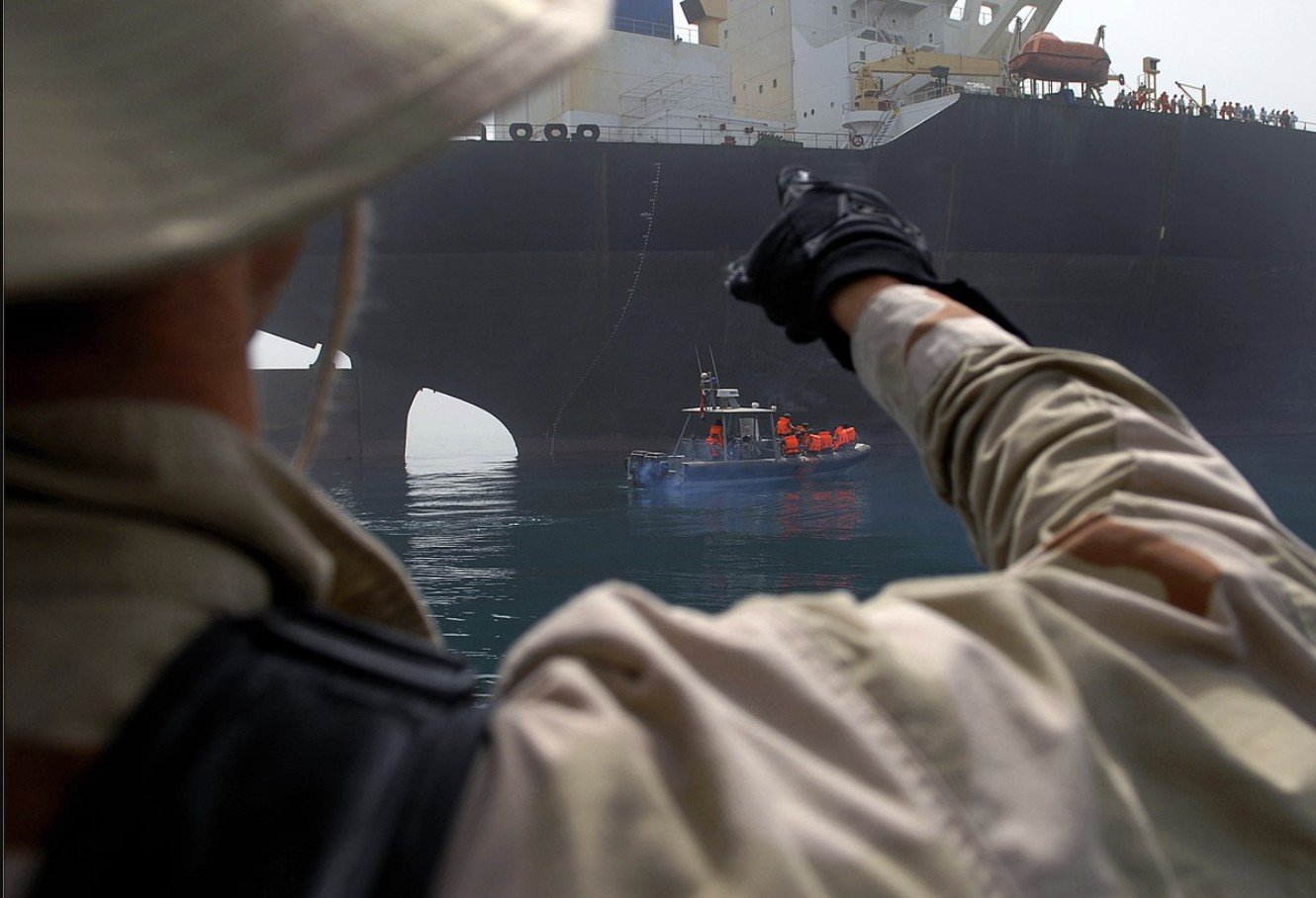 A crew member points towards a large cargo ship while a smaller boat approaches, showcasing maritime operations and teamwork in a busy harbor.