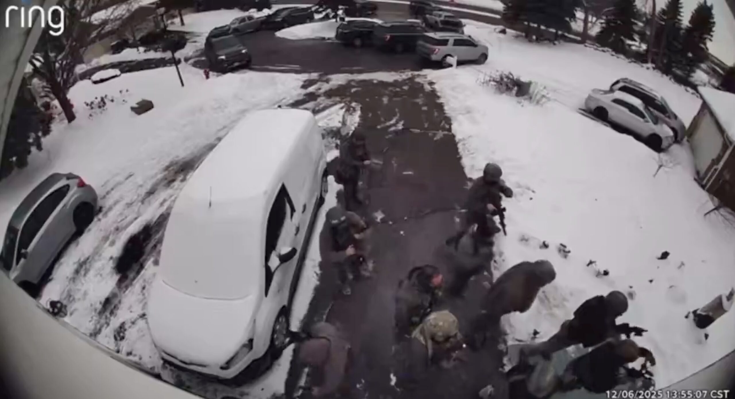 Law enforcement officers in tactical gear approach a residential driveway covered in snow, with parked vehicles visible in the background.