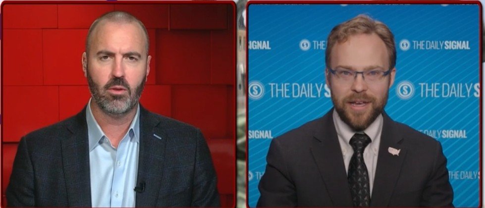 Two men engage in a discussion, with one in a blazer and the other in a suit, set against a backdrop featuring The Daily Signal logo.