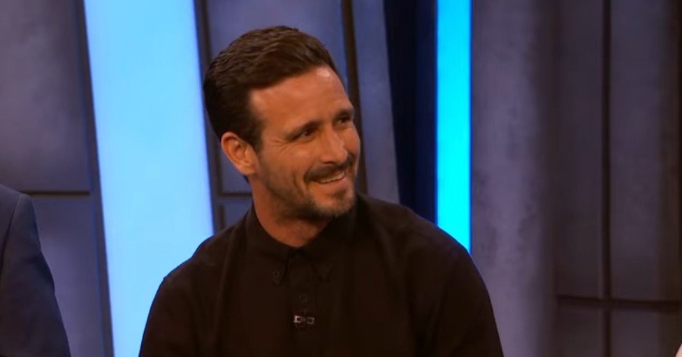 Smiling man in a black shirt sitting on a talk show set with blue lighting in the background.