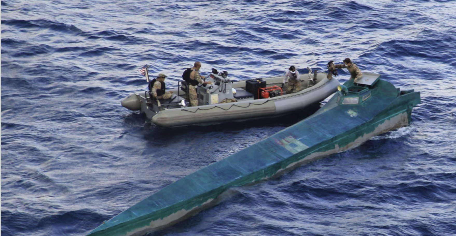 Military personnel inspect a semi-submersible vessel while aboard a small boat in open ocean waters.