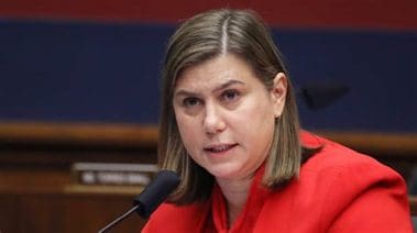 Woman in a red blazer speaking at a congressional hearing, focused on addressing key issues with a microphone in front of her.