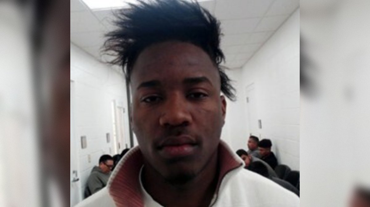Mugshot of a young man with distinctive hairstyle, sitting in a waiting area with others in the background.