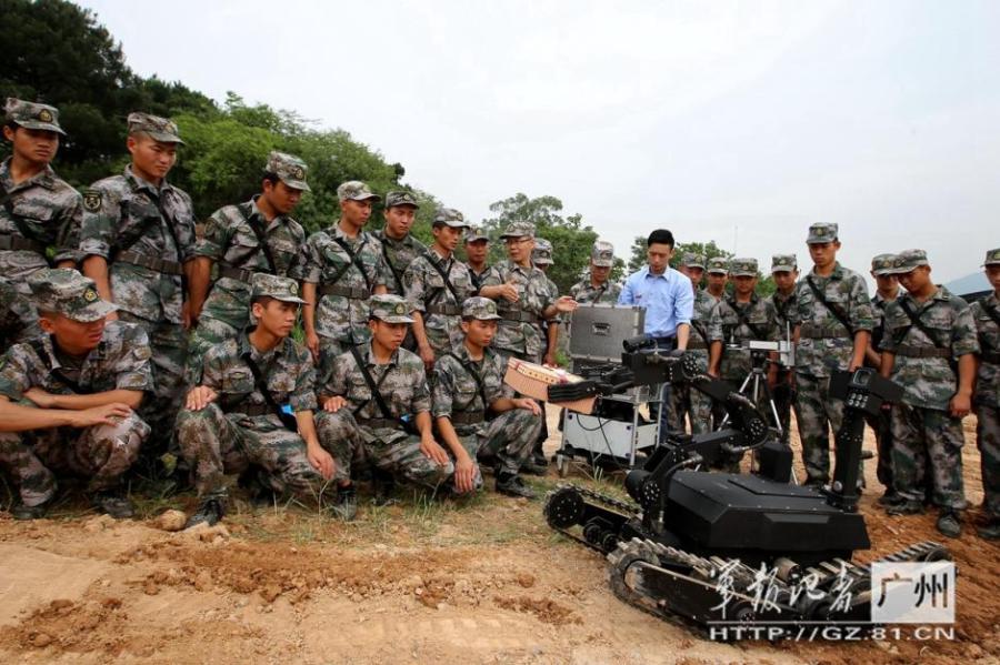 Military personnel observe a demonstration of a robotic device during a training exercise in a rural setting.