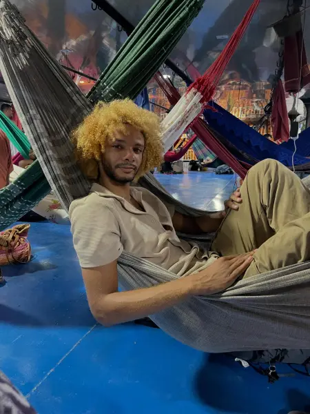 Young man with curly blonde hair relaxing in a hammock in a vibrant indoor space filled with colorful hammocks.