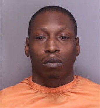 Mugshot of a man wearing an orange jumpsuit, facing the camera with a neutral expression, against a gray background.