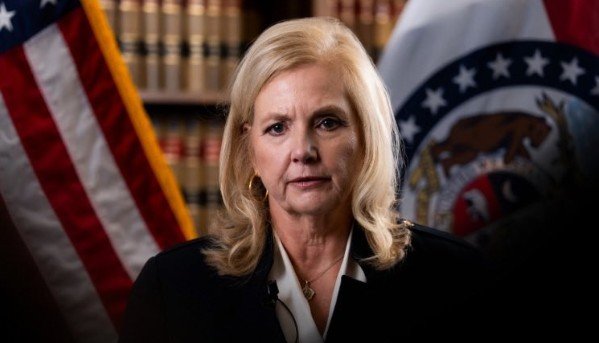 Woman in a black suit stands in front of American and Missouri state flags, speaking in a formal setting with bookshelves in the background.