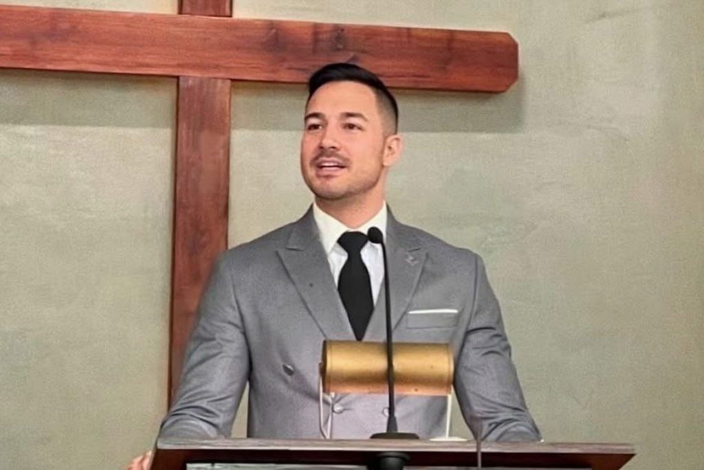Man in a gray suit delivering a speech at a podium in front of a wooden cross in a church setting.