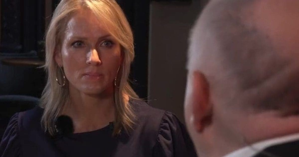A woman with blonde hair and earrings is engaged in a serious conversation with a man, focusing intently on the discussion in a dimly lit setting.