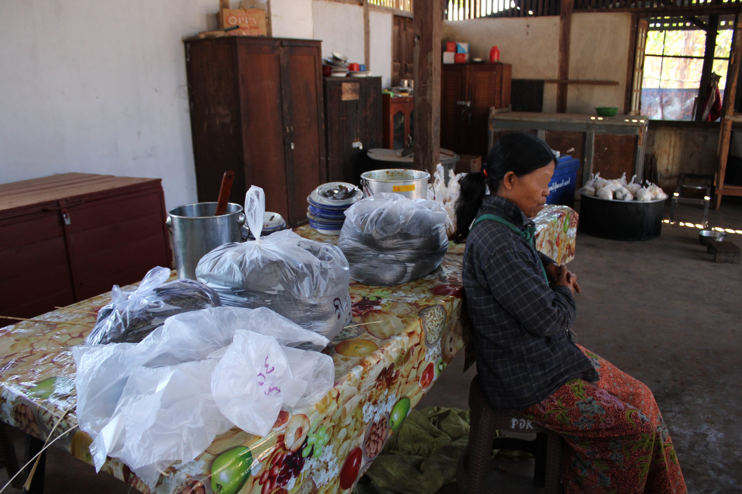 Woman sitting at a table in a rustic kitchen, surrounded by bags of food and cooking utensils, highlighting traditional food preparation practices.