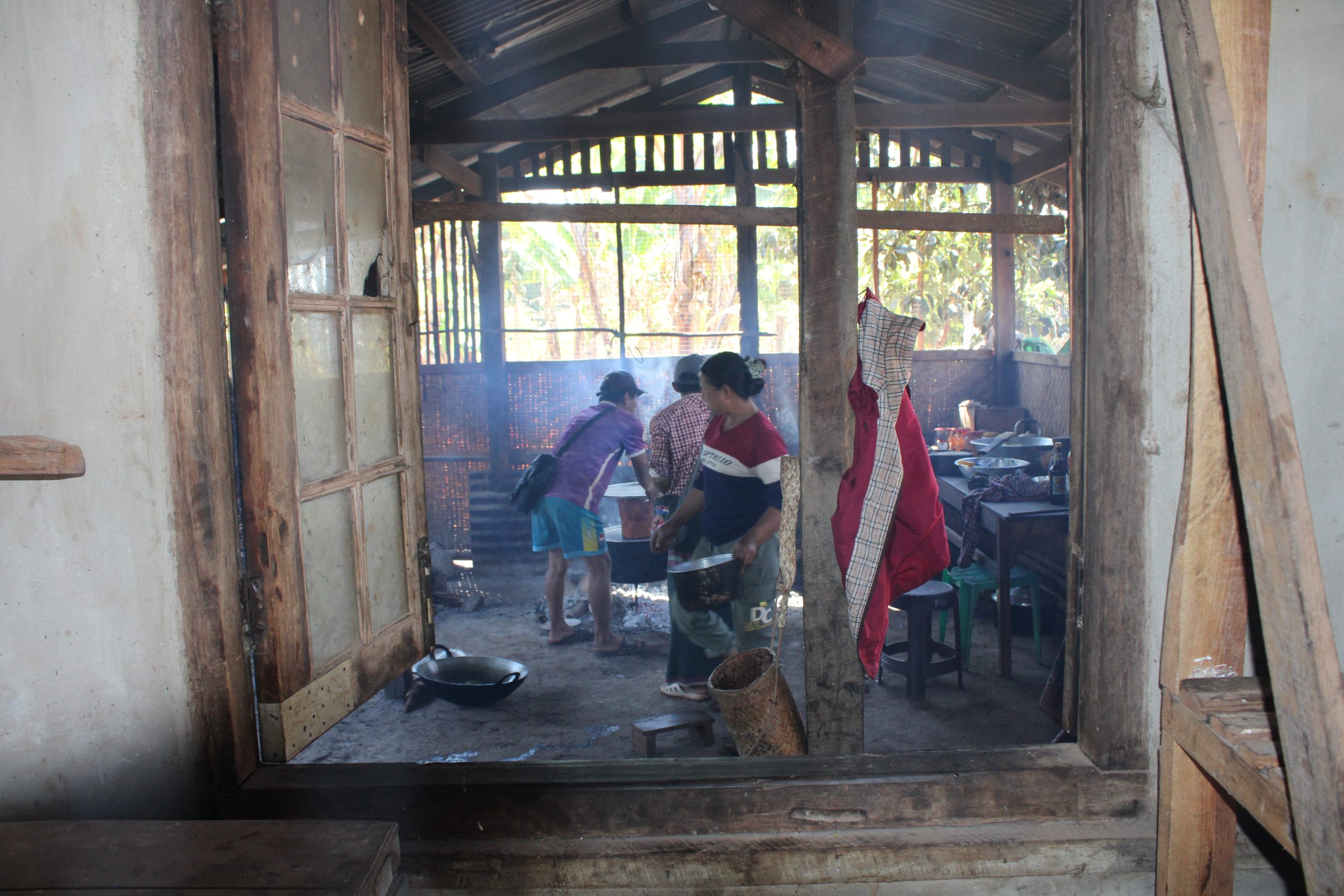 Interior view of a rustic kitchen with two women cooking, surrounded by wooden structures and smoke, capturing a traditional cooking scene.