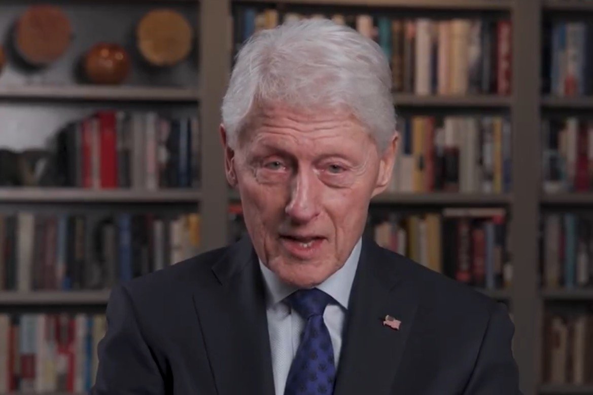 Former President Bill Clinton speaks on various topics, with a bookshelf filled with books and decorative items in the background.