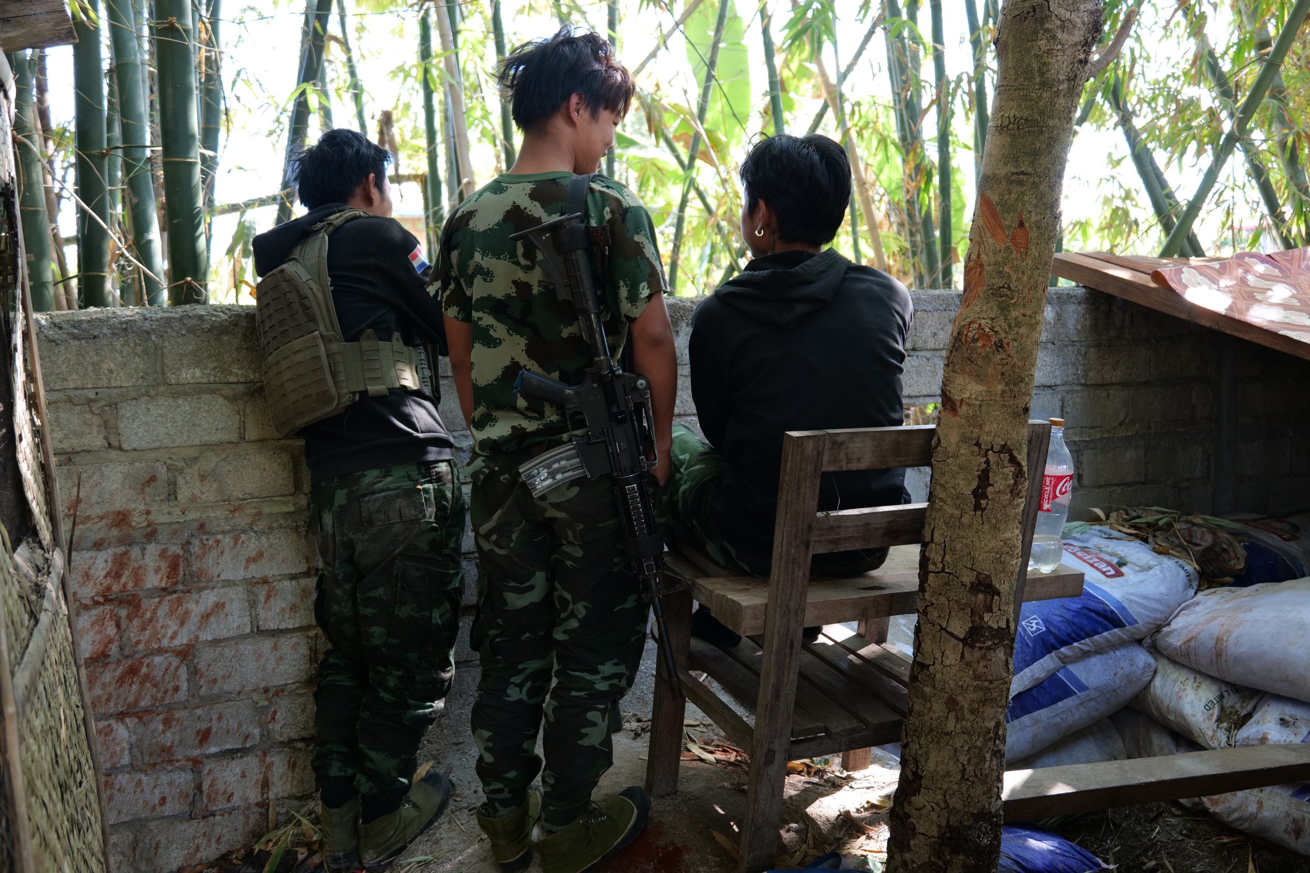 Three individuals in camouflage attire are gathered near a stone wall in a wooded area, with one person seated on a wooden stool.