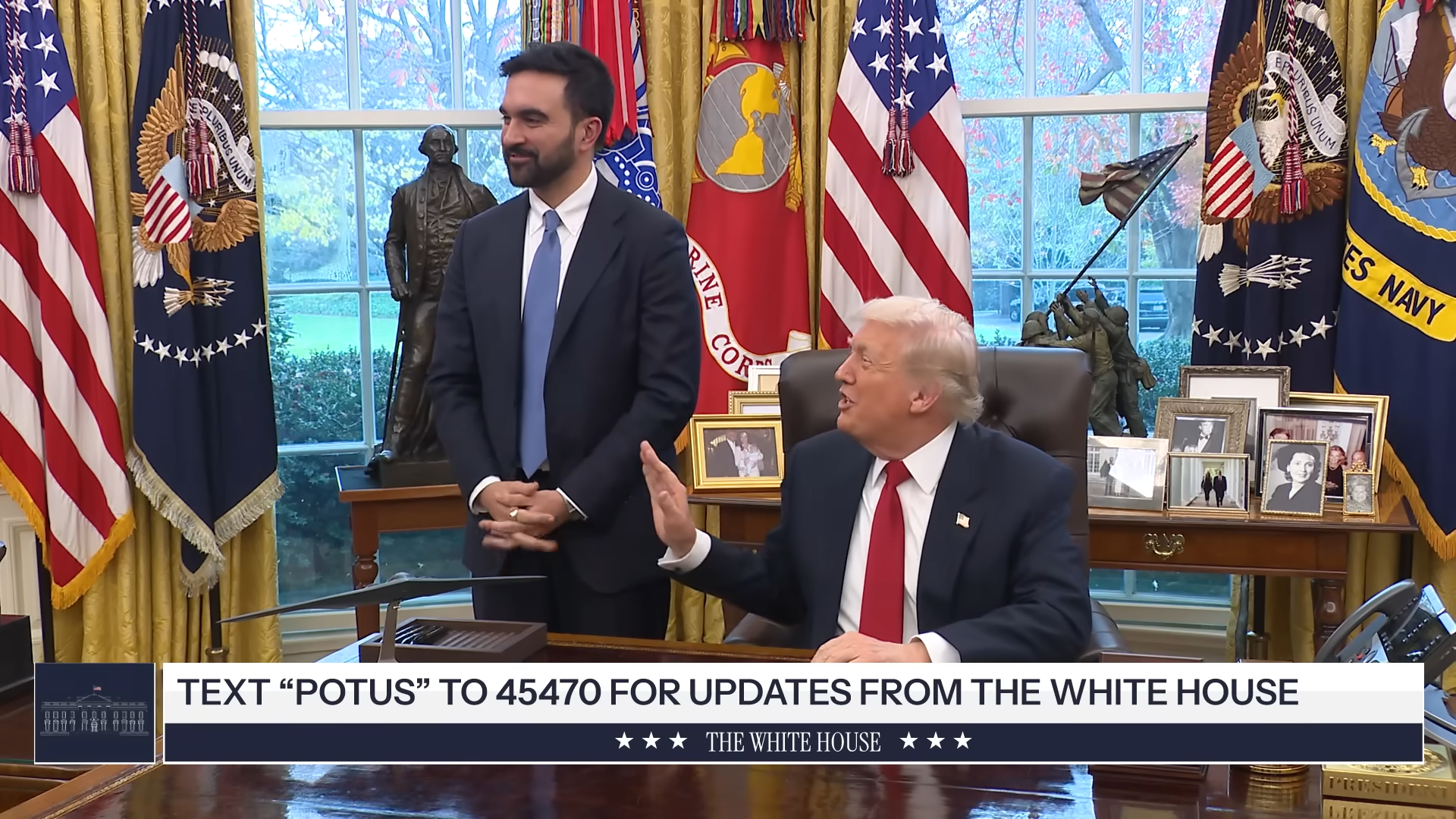 Donald Trump speaks while a staff member stands beside him in the Oval Office, surrounded by U.S. flags and presidential memorabilia.