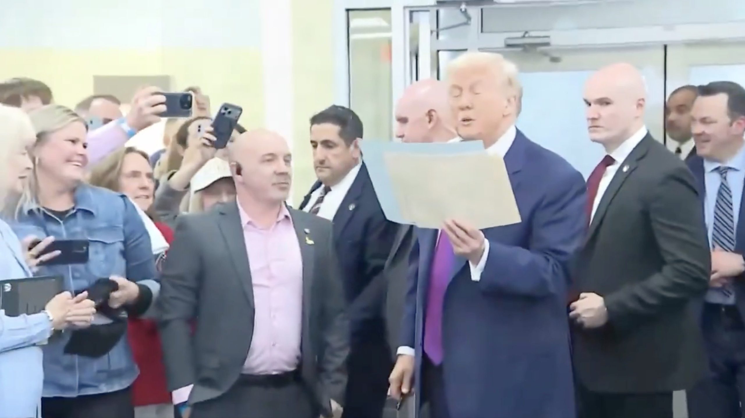 Donald Trump speaks to a crowd while holding a document, surrounded by supporters and security personnel at a public event.