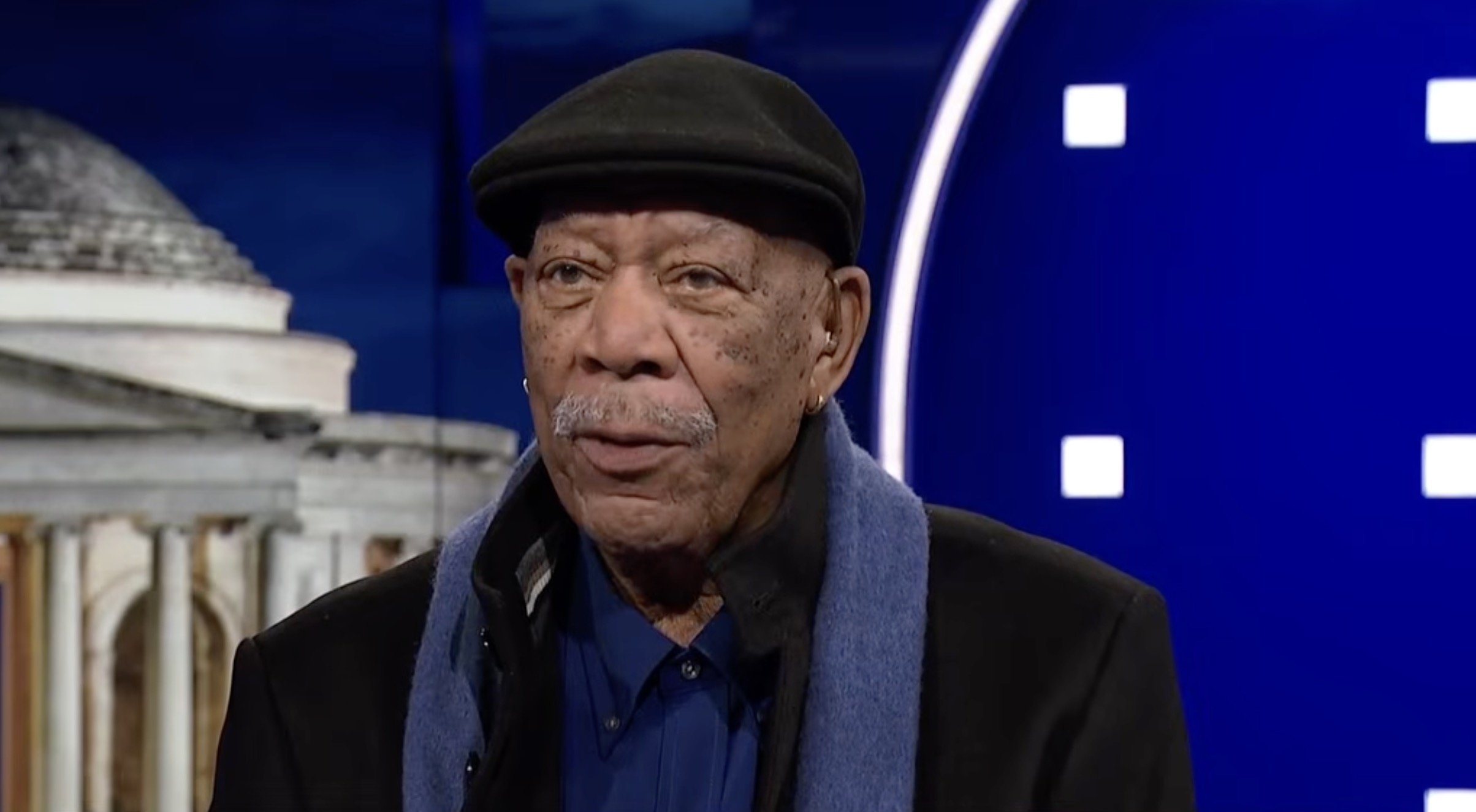 Elderly man wearing a black cap and blue scarf speaks during an interview on a television set with a blue background.
