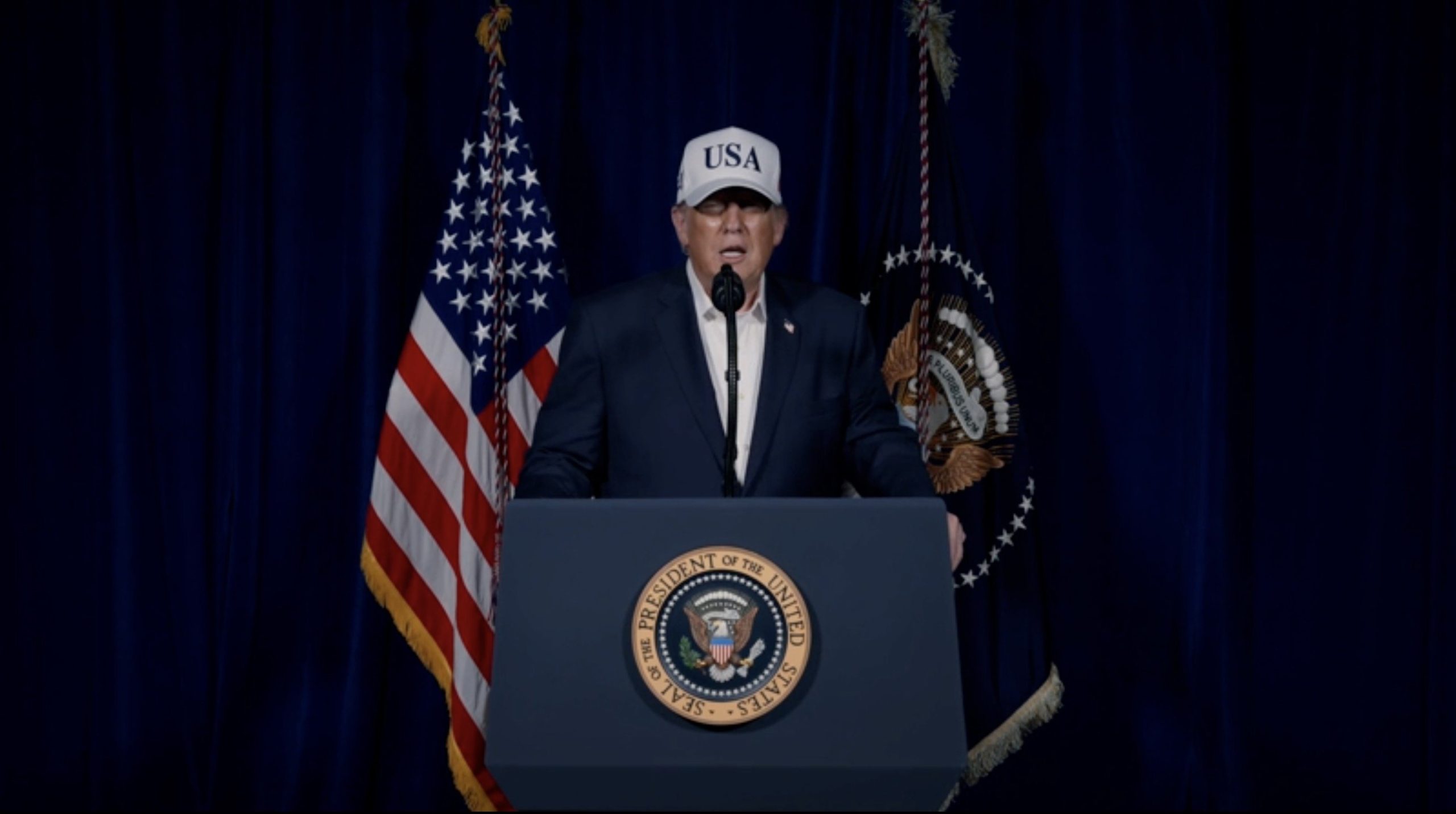 Former President speaking at a podium with the presidential seal, flanked by U.S. flags, wearing a cap that says "USA."
