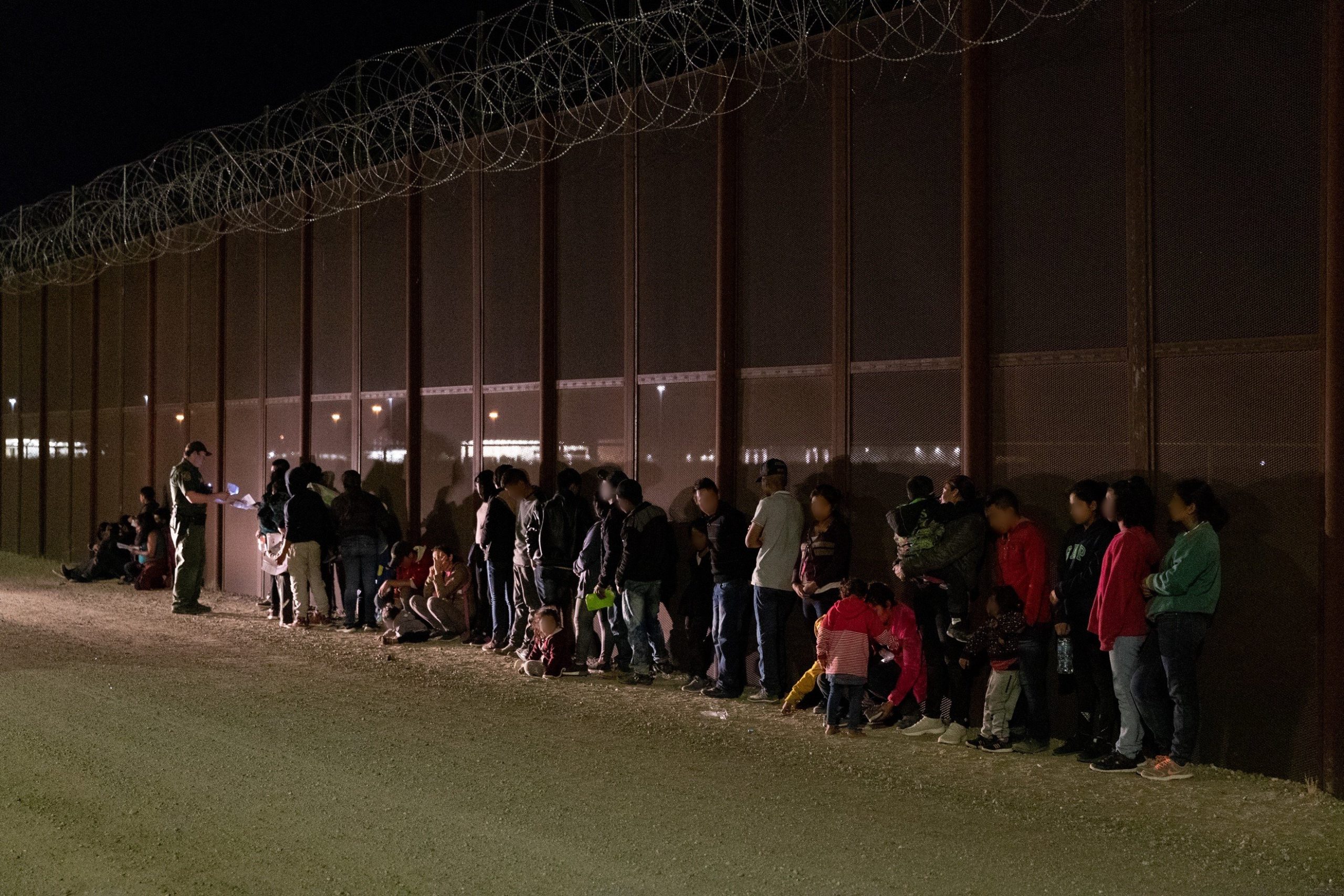 A line of people waits near a border wall at night, with a border patrol agent checking documents under artificial lighting.