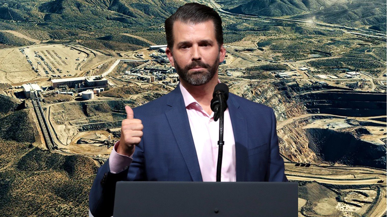 Donald Trump Jr. giving a thumbs up during a speech at a mining site with mountainous terrain in the background.