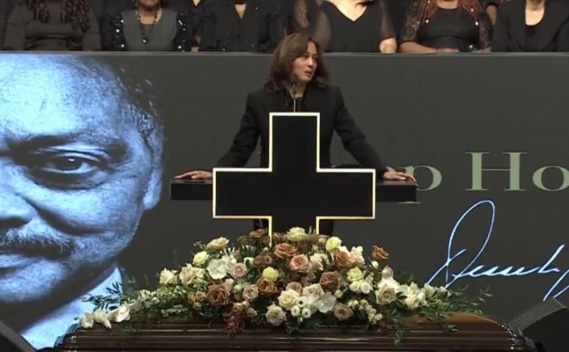 Speaker delivering a eulogy at a memorial service, with a large floral arrangement and a prominent cross in the foreground, honoring a notable figure.