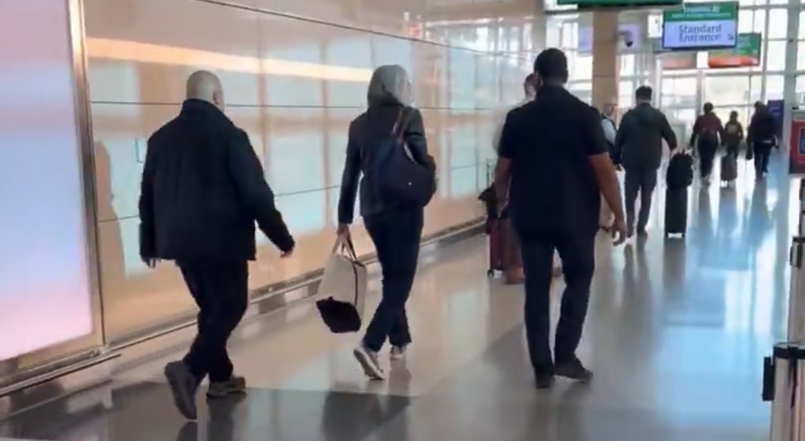 People walking through a busy airport terminal, with luggage and signs in the background.