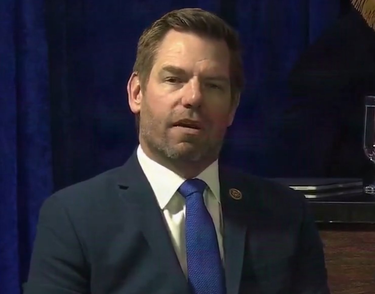 Man in a suit with a blue tie, seated against a blue backdrop, appearing thoughtful during a formal event.