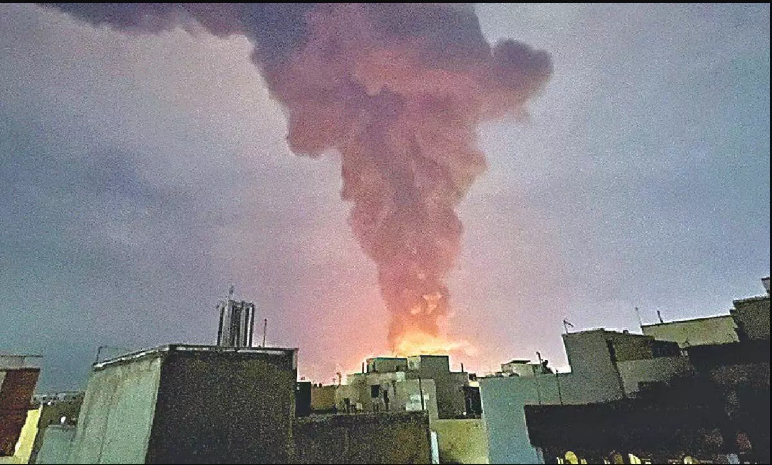 Thick smoke billows from a cityscape at dusk, indicating a possible fire or explosion in the area.