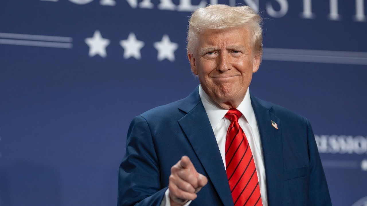 Donald Trump smiles and points while wearing a blue suit and red tie at a public event, with a backdrop featuring stars and text.