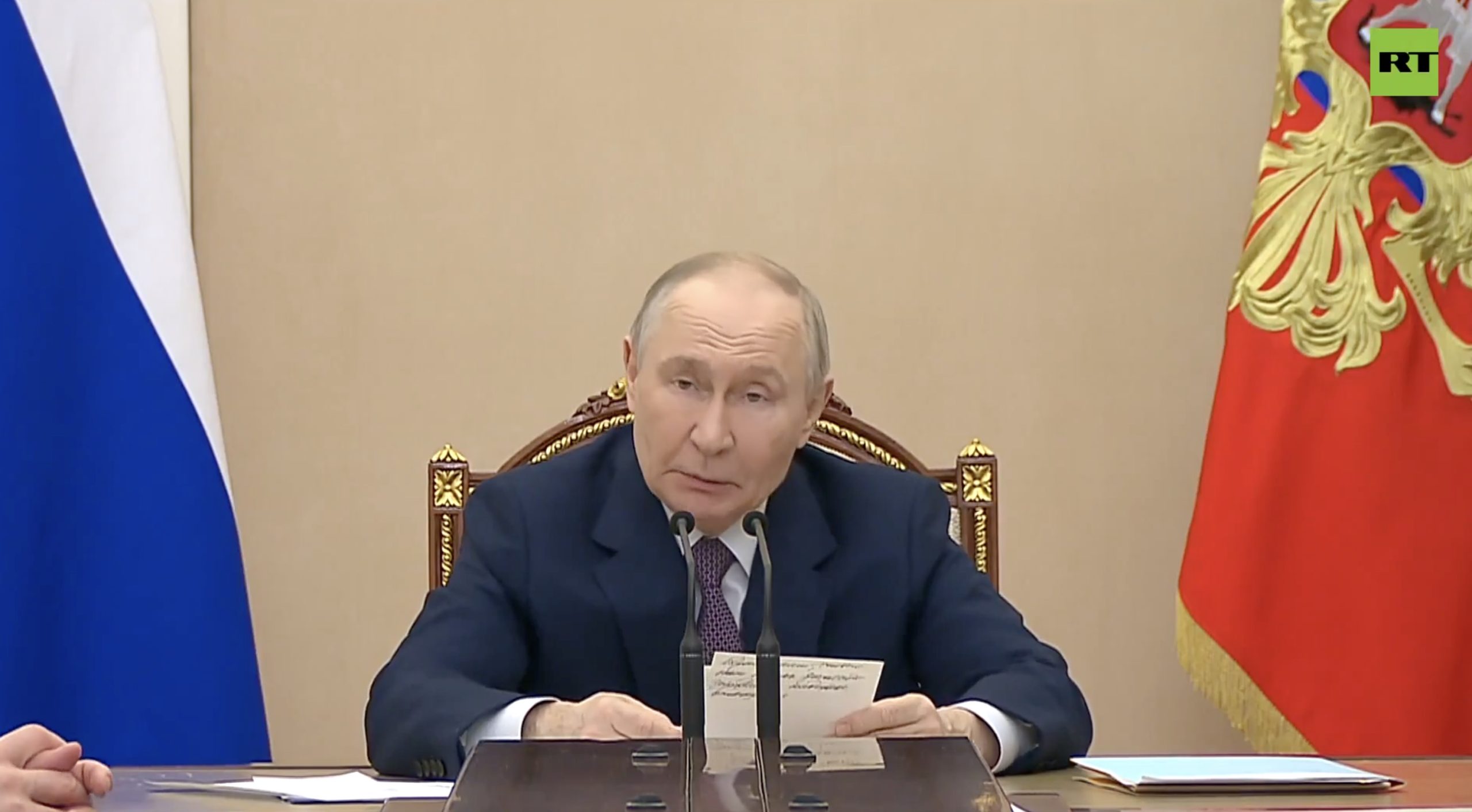 Russian leader addressing officials during a meeting, with the Russian flag and coat of arms in the background.