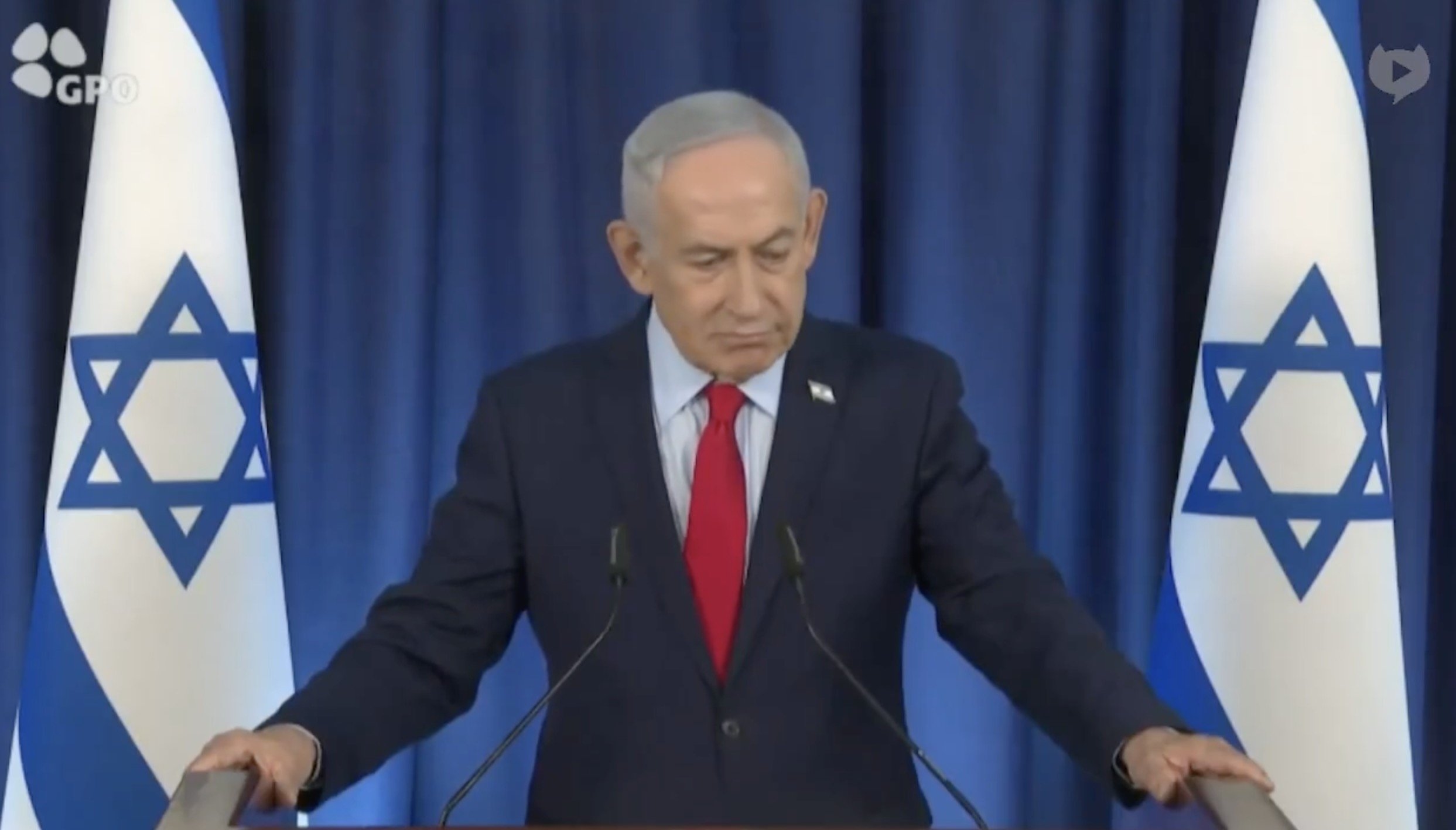 Israeli Prime Minister Benjamin Netanyahu speaks at a podium with Israeli flags in the background during an official address.