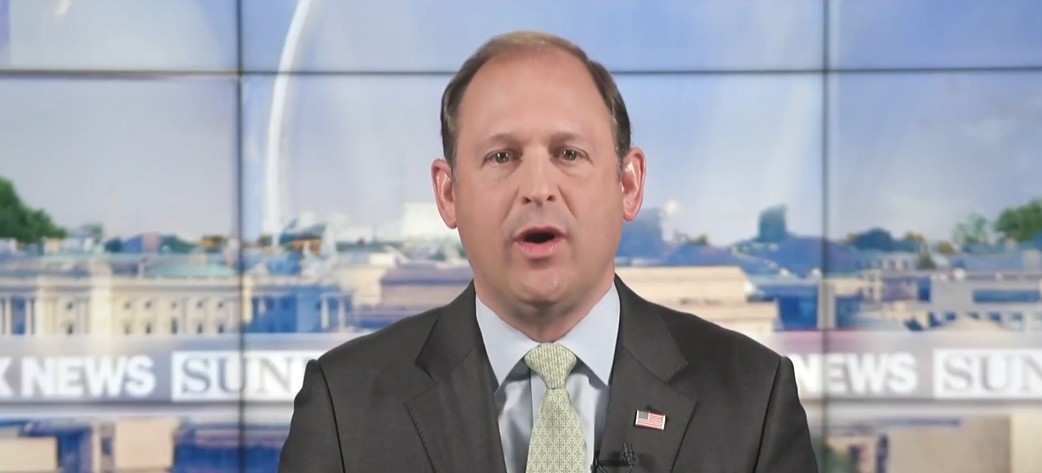 News anchor speaking on a televised segment with a backdrop of the White House and a clear blue sky.