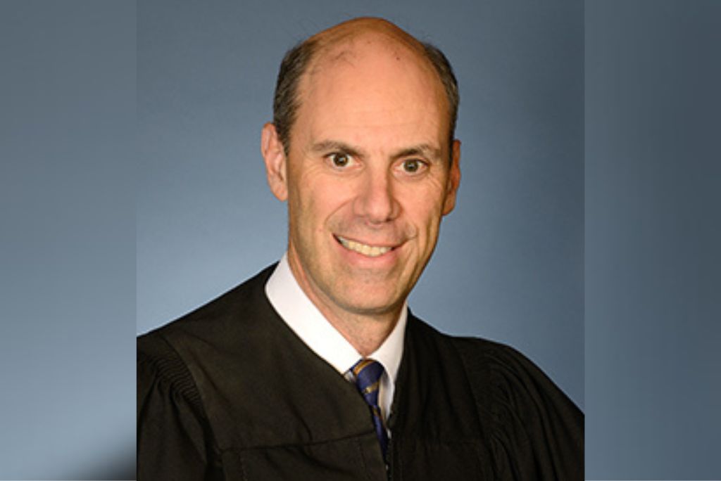 Portrait of a judge in a black robe, smiling against a blue background, representing the judicial profession and authority in law.