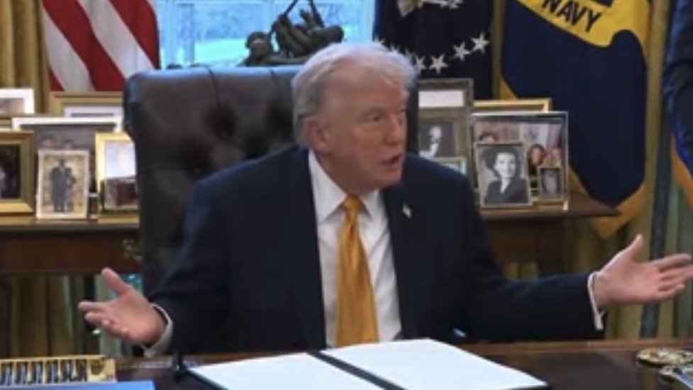 Former President Donald Trump gestures while speaking in the Oval Office, surrounded by framed photos and official seals.