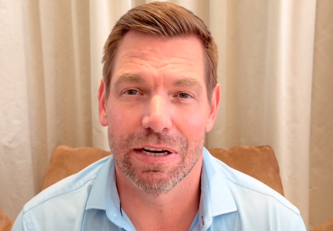 Smiling man in a light blue shirt sitting in front of a beige curtain, speaking directly to the camera in a casual setting.