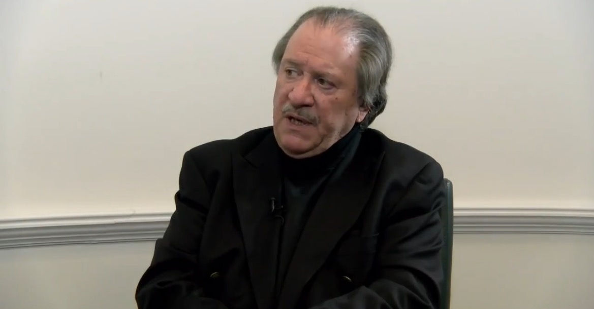 Man in a black jacket sitting in an interview setting, engaged in conversation with a serious expression against a neutral background.
