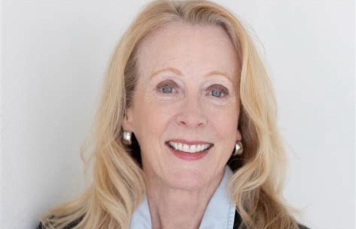 Smiling woman with long blonde hair and earrings, wearing a black jacket and light blue shirt against a neutral background.