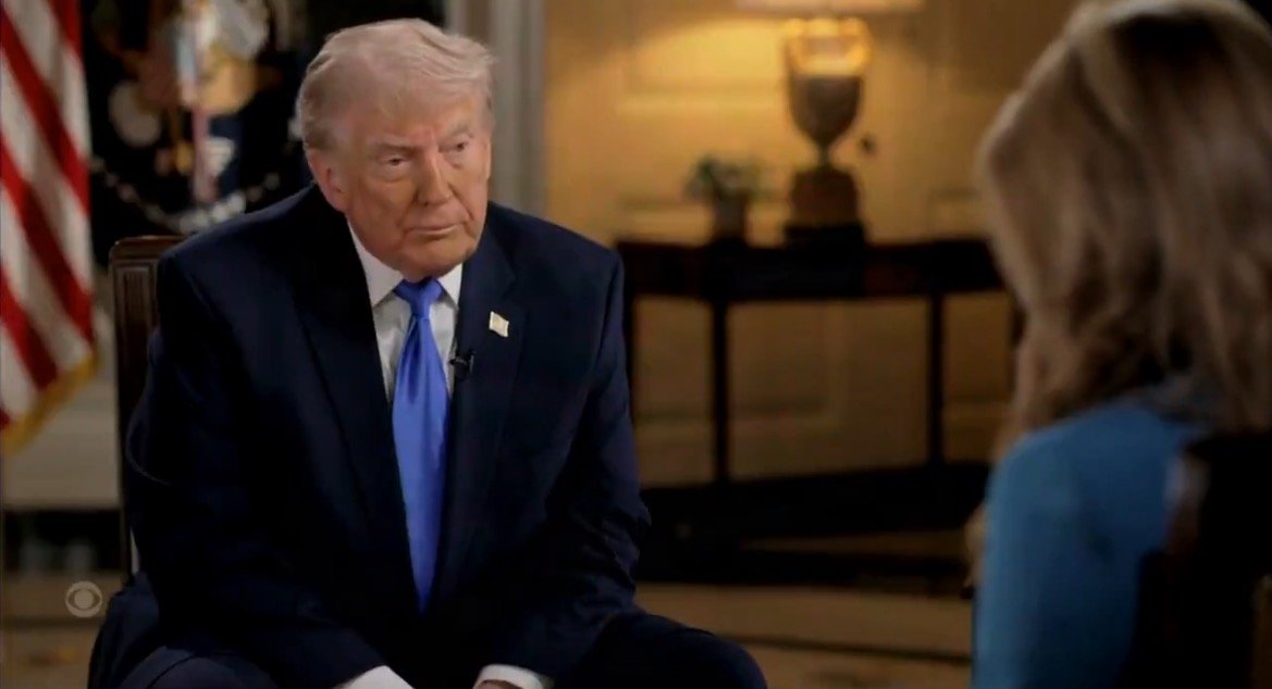 Donald Trump sits for an interview in a formal setting, wearing a dark suit and blue tie, with an interviewer partially visible in the foreground.