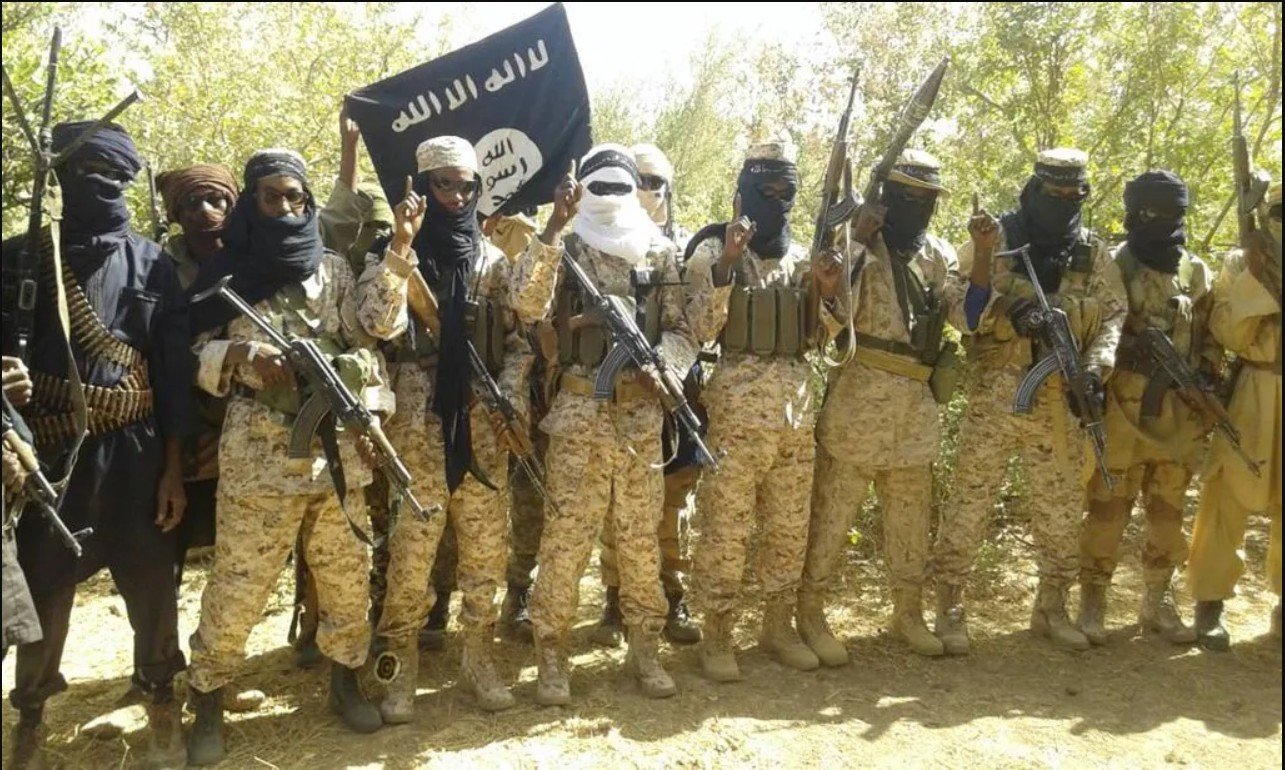 Militant group members posing with weapons and a black flag in a forested area, showcasing military attire and a display of allegiance.