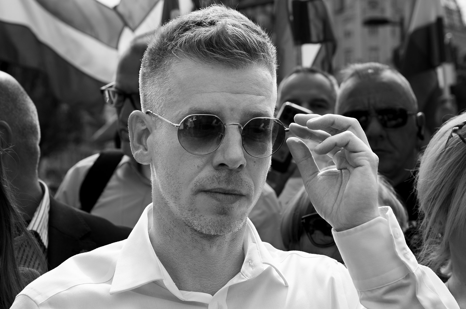 Man in sunglasses adjusting his glasses at a crowded event with flags in the background, captured in black and white.
