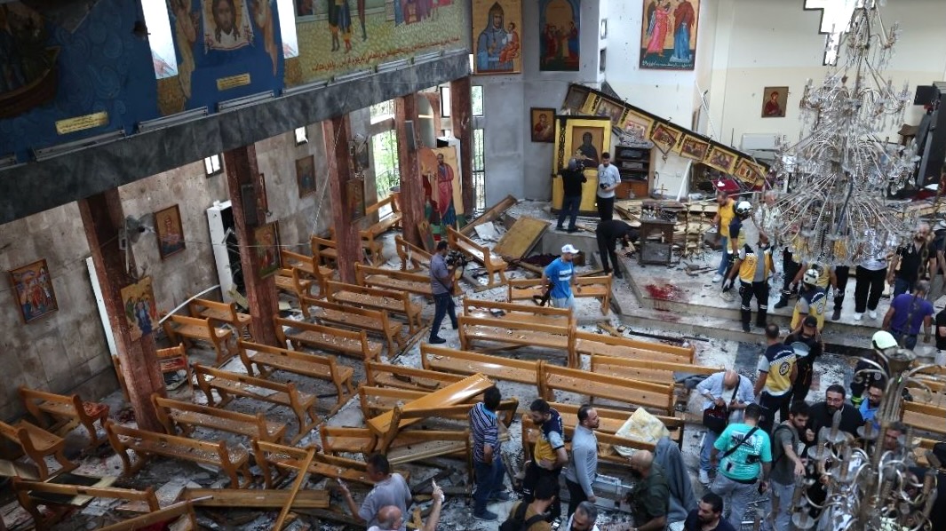 Interior of a damaged church with scattered benches and debris, showing people assessing the destruction after an incident.